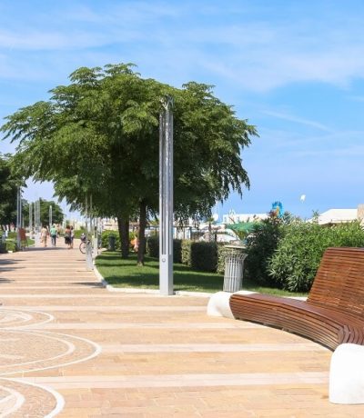 Passeggiata lungo un viale pedonale con panchine moderne, alberi verdi e cielo sereno.