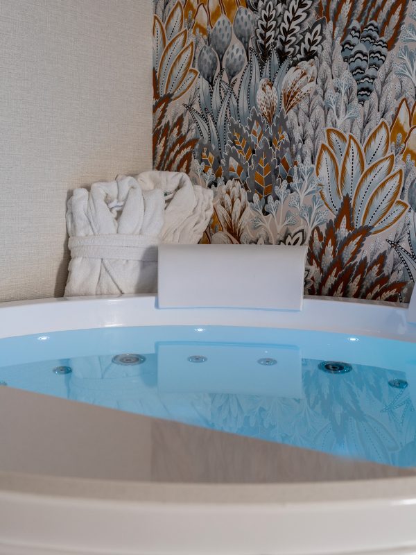 A round bathtub filled with water, next to a white robe and colorful patterned wallpaper.