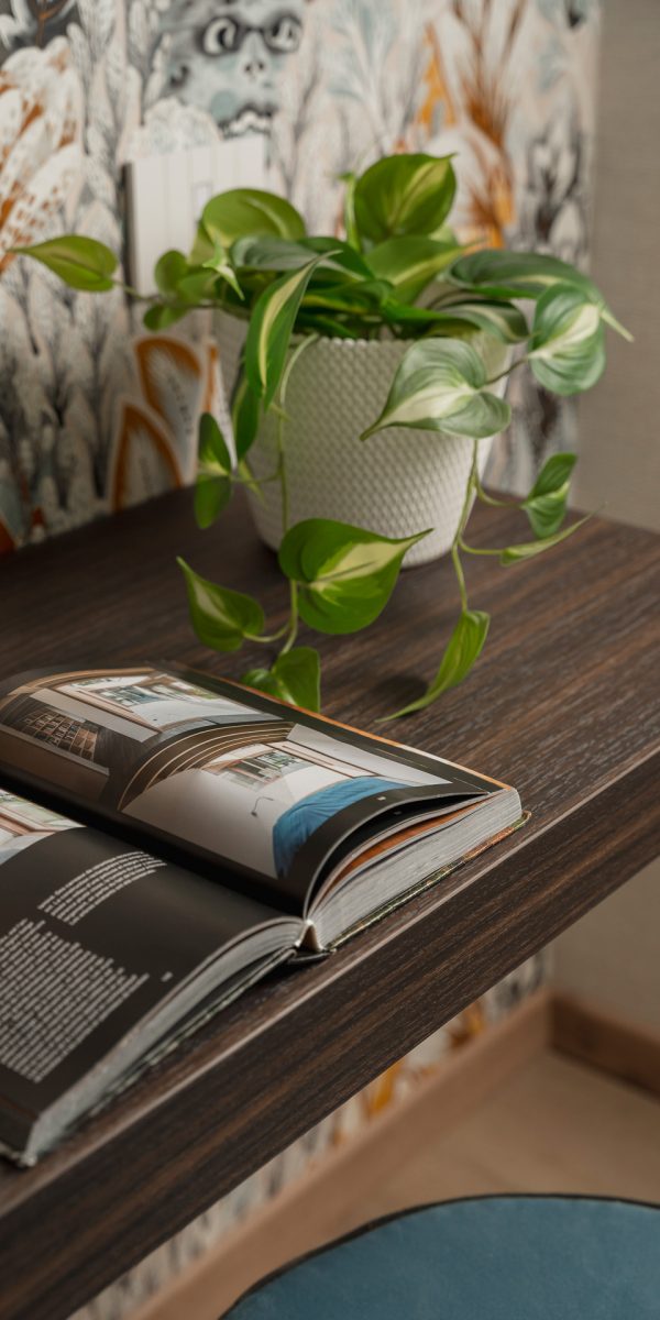 Open book and potted plant on a wooden table with patterned wallpaper in the background.