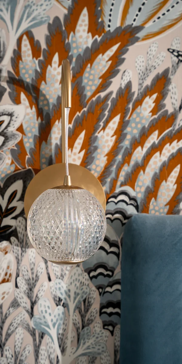 A crystal wall sconce with a golden base against a colorful floral-patterned wallpaper.
