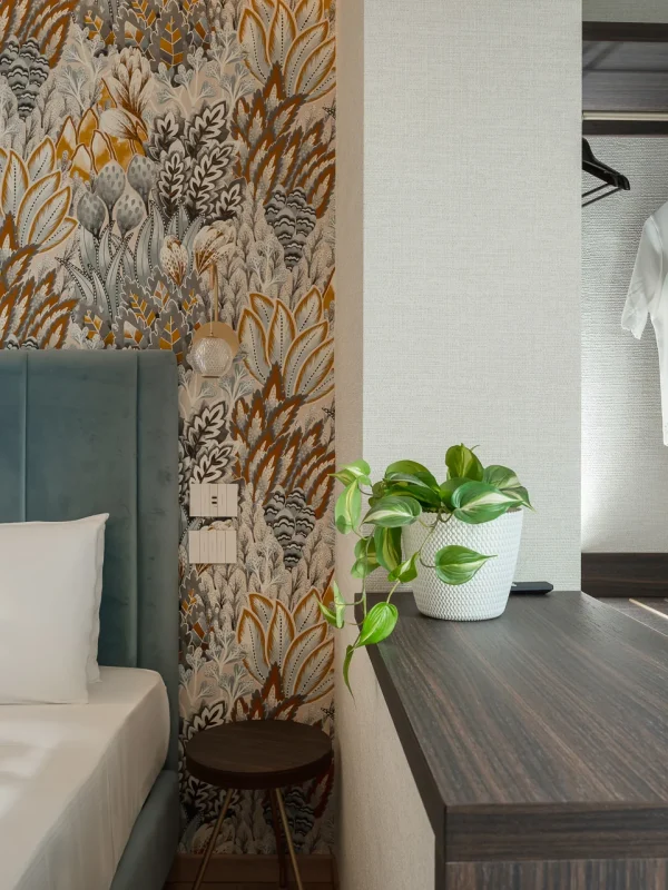 A cozy bedroom with a patterned wall, a plush bed, a plant on a wooden desk, and clothes hanging in an open closet.